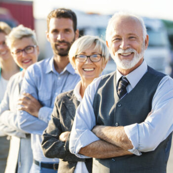 Closeup front view of multi aged corporate team standing in a row in front of their company. There are many semi trucks in background. Senior executive is in front, two more men and three women behind him.